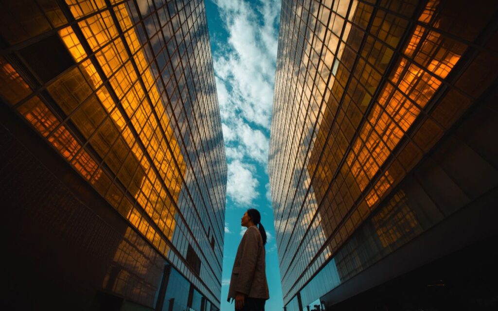 View from ground towards sky with person standing in the gap between symmetrical modern glass buildings.